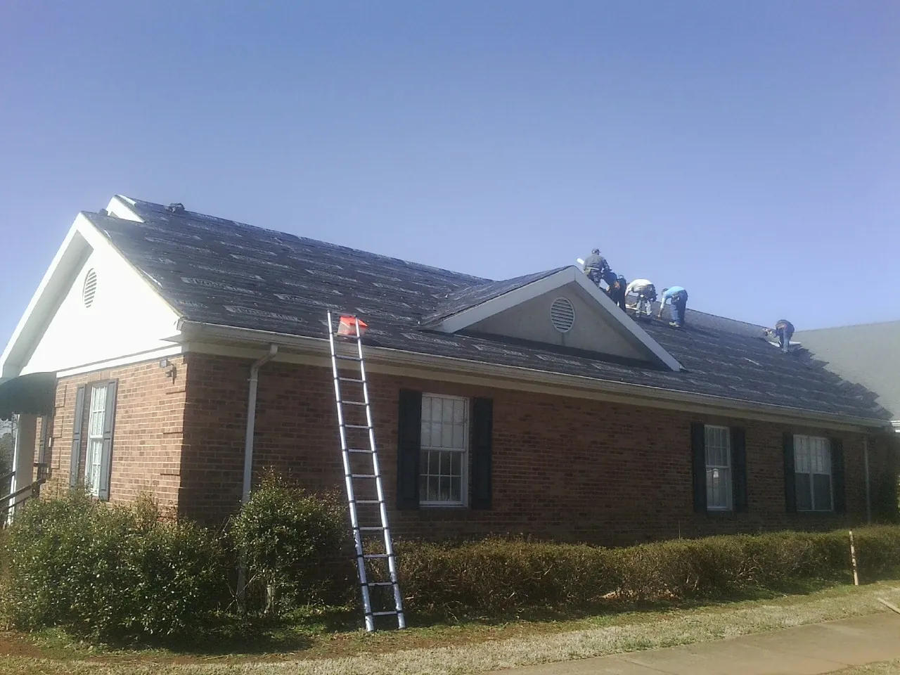 Crew tearing shingles off a brick ranch roof with a ladder set — Emergency Roof Repair work by our crew in Momence