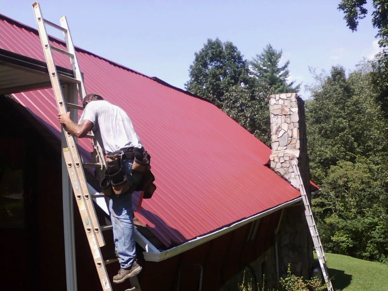 Roofer on a ladder beside a red metal roof and stone chimney — Soffit & Fascia Repair by Roofing Brothers in Momence